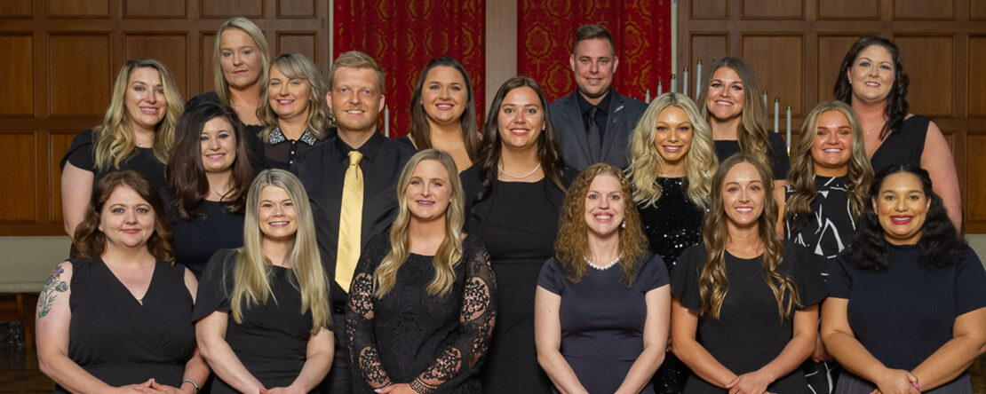 A group of nineteen people, mostly women and one man, dressed in black attire, stand together in three rows, smiling at the camera in a formal indoor setting with wood paneling and red curtains.