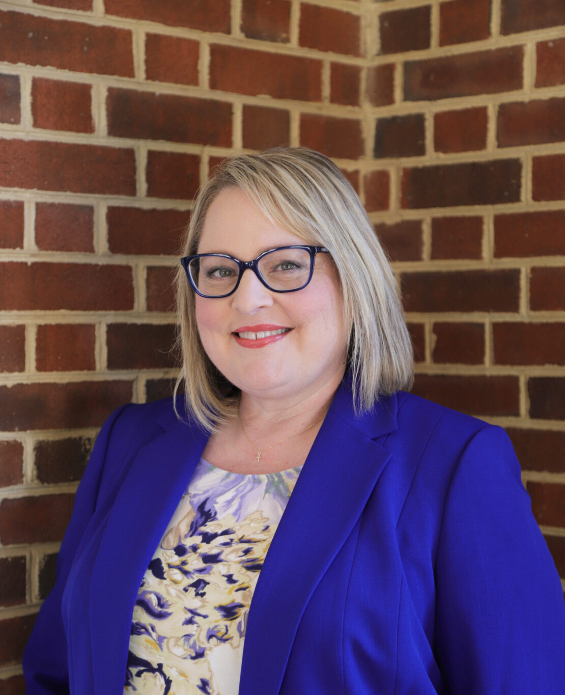 A woman with blonde hair and glasses smiles, wearing a blue blazer and a floral blouse, standing in front of a red brick wall.