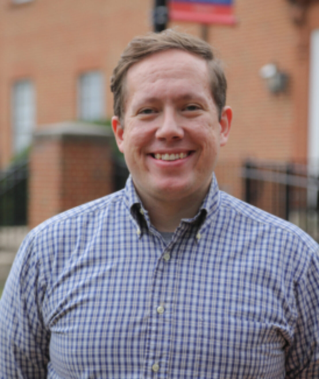 A smiling man wearing a blue and white plaid shirt stands outdoors in front of a brick building with blurred signage and windows in the background.