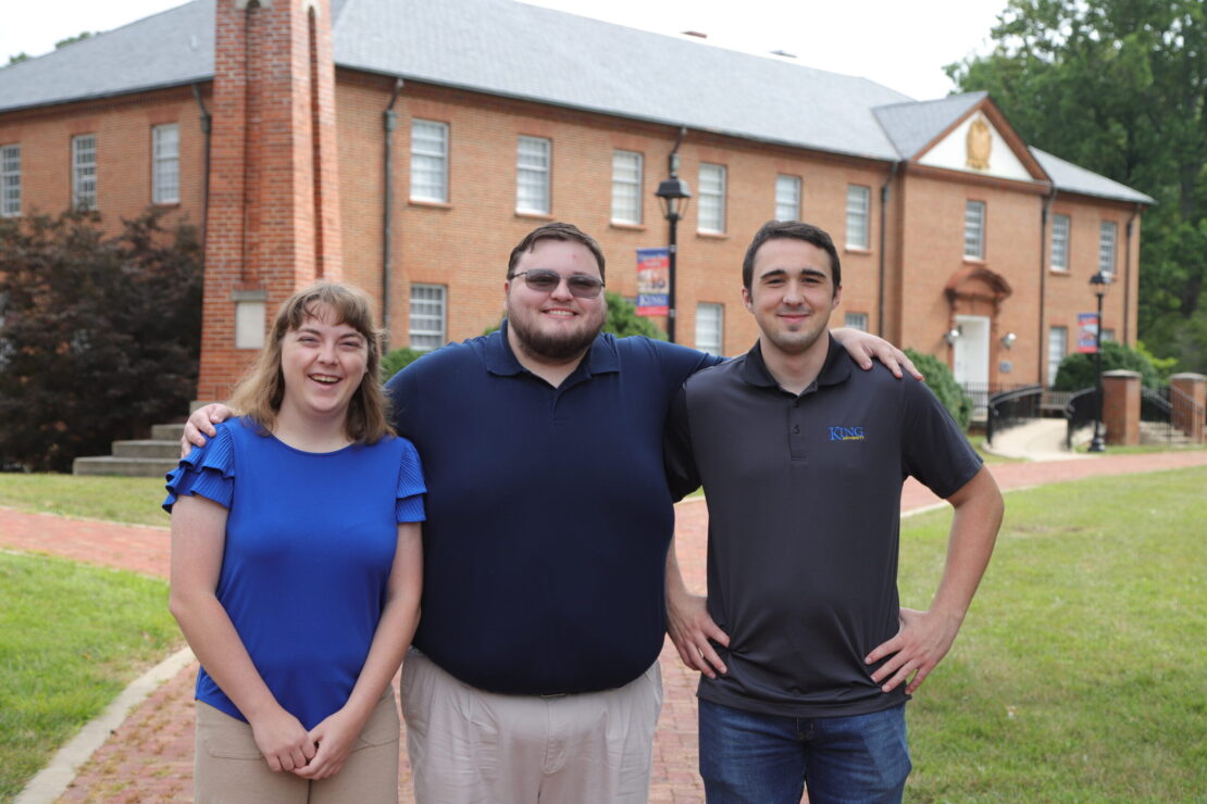 Three people stand outside on a campus, smiling at the camera. The two men and one woman are dressed casually, with a brick building and green grass in the background.