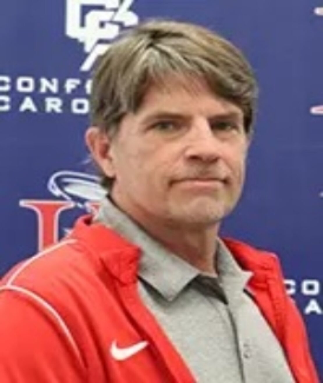 A middle-aged man with short brown hair wearing a gray collared shirt and a red jacket, standing in front of a blue background with logos and text.