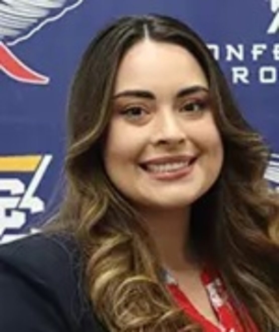 A woman with long wavy brown hair, wearing a dark blazer over a red and white top, smiles in front of a blue background with sports logos and white text.