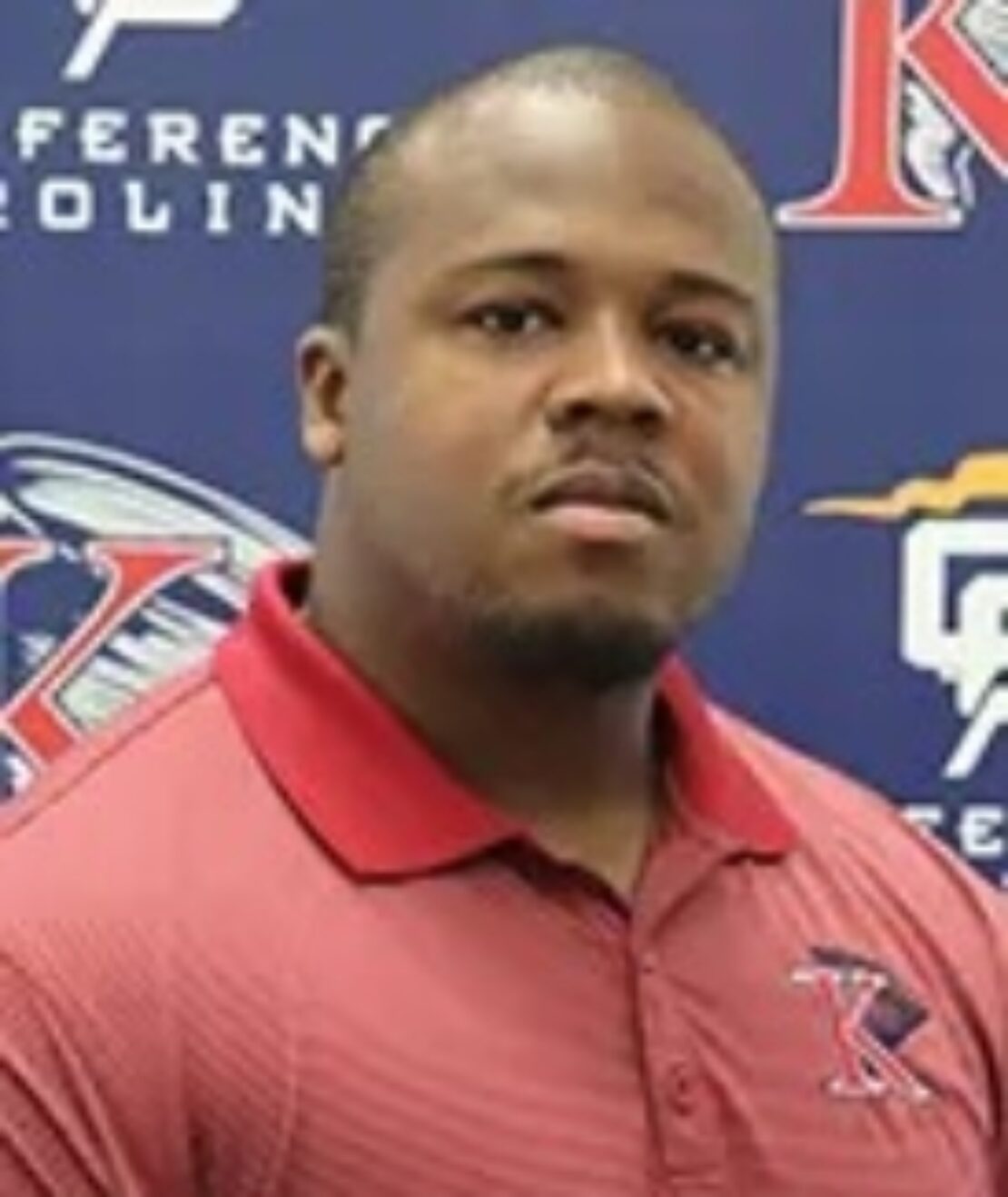 A man wearing a red polo shirt stands in front of a blue backdrop featuring sports team logos and text. He looks directly at the camera with a neutral expression.