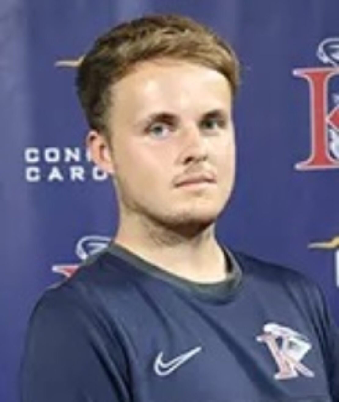 A young man with short light brown hair and a trimmed beard, wearing a navy sports jersey with a K logo, stands in front of a blue backdrop with logos and text.
