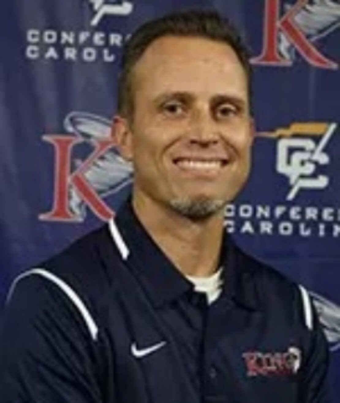 A man with short hair and a goatee smiles while wearing a dark polo shirt with a King logo. The background features Conference Carolinas and King University logos.