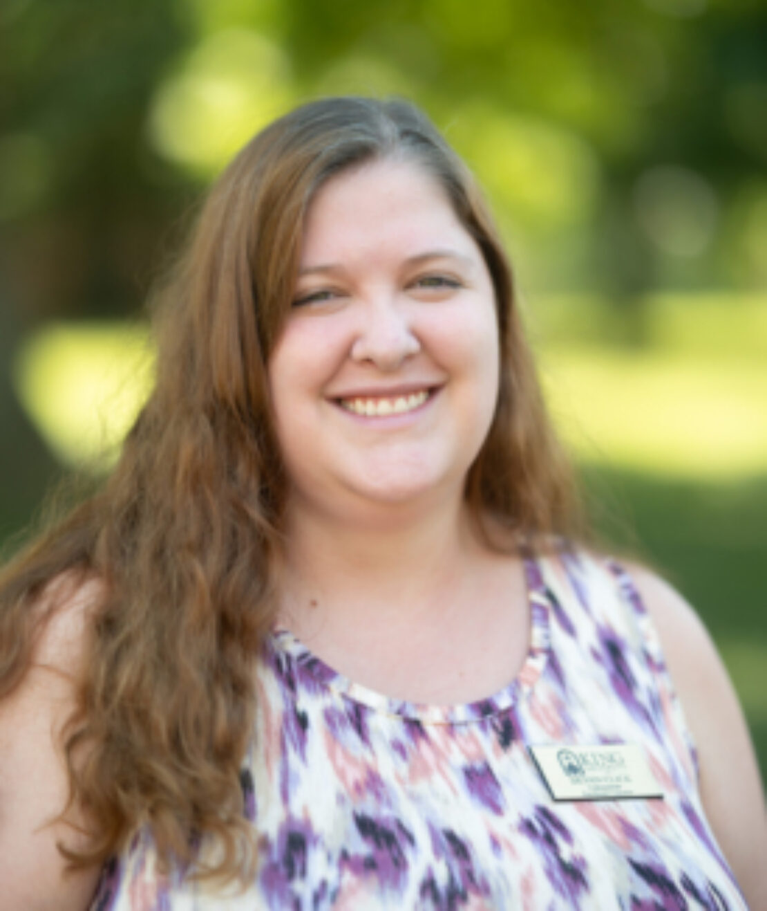 A woman with long, wavy brown hair smiles outdoors. She is wearing a sleeveless patterned top and a name tag. The background is blurred greenery.