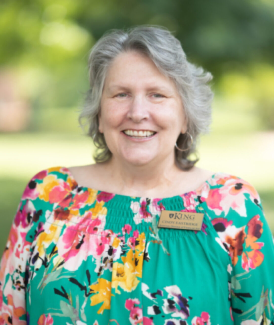 A smiling older woman with gray hair wears a colorful floral blouse and a name tag reading KING and Linda Easterling. She stands outdoors with trees and greenery blurred in the background.