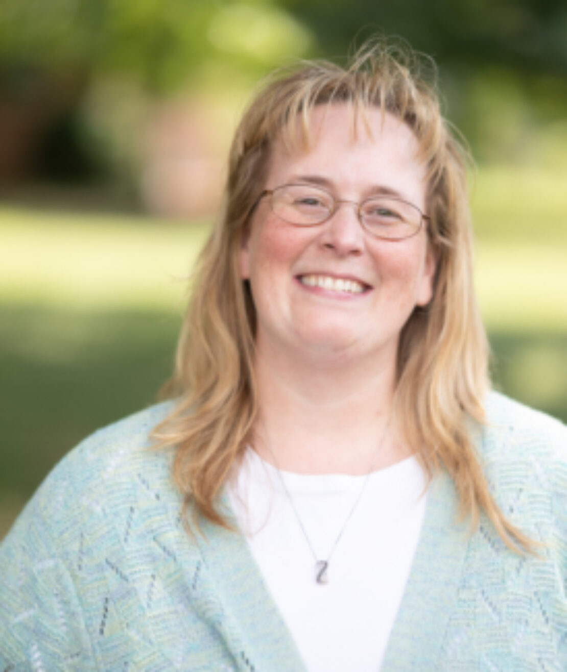 A woman with blonde hair, glasses, and a light blue patterned cardigan over a white shirt smiles outdoors with a blurred green background.