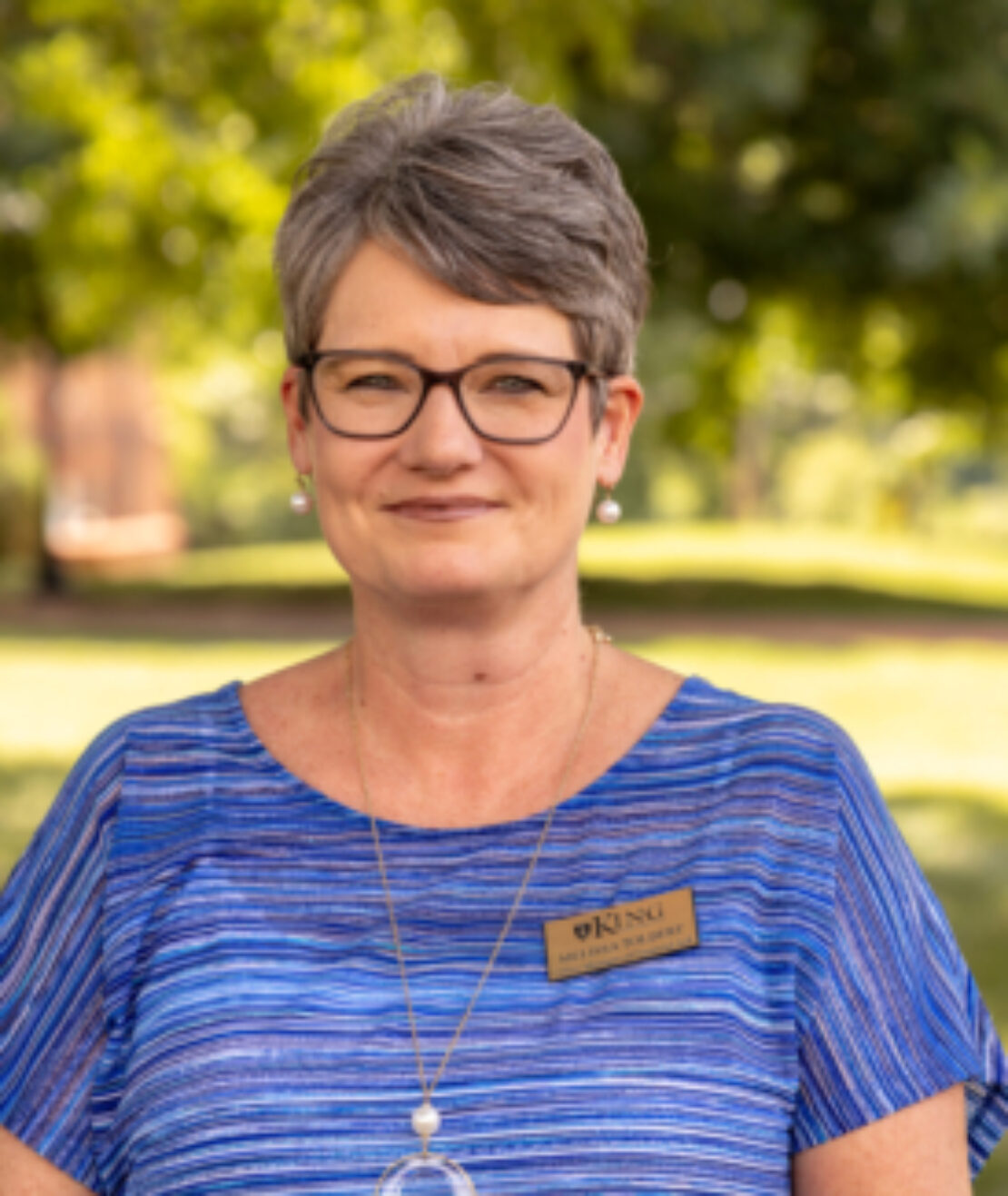 A middle-aged woman with short gray hair and glasses wears a blue striped blouse, a pendant necklace, and a name tag that reads KING. She stands outdoors with green trees blurred in the background.