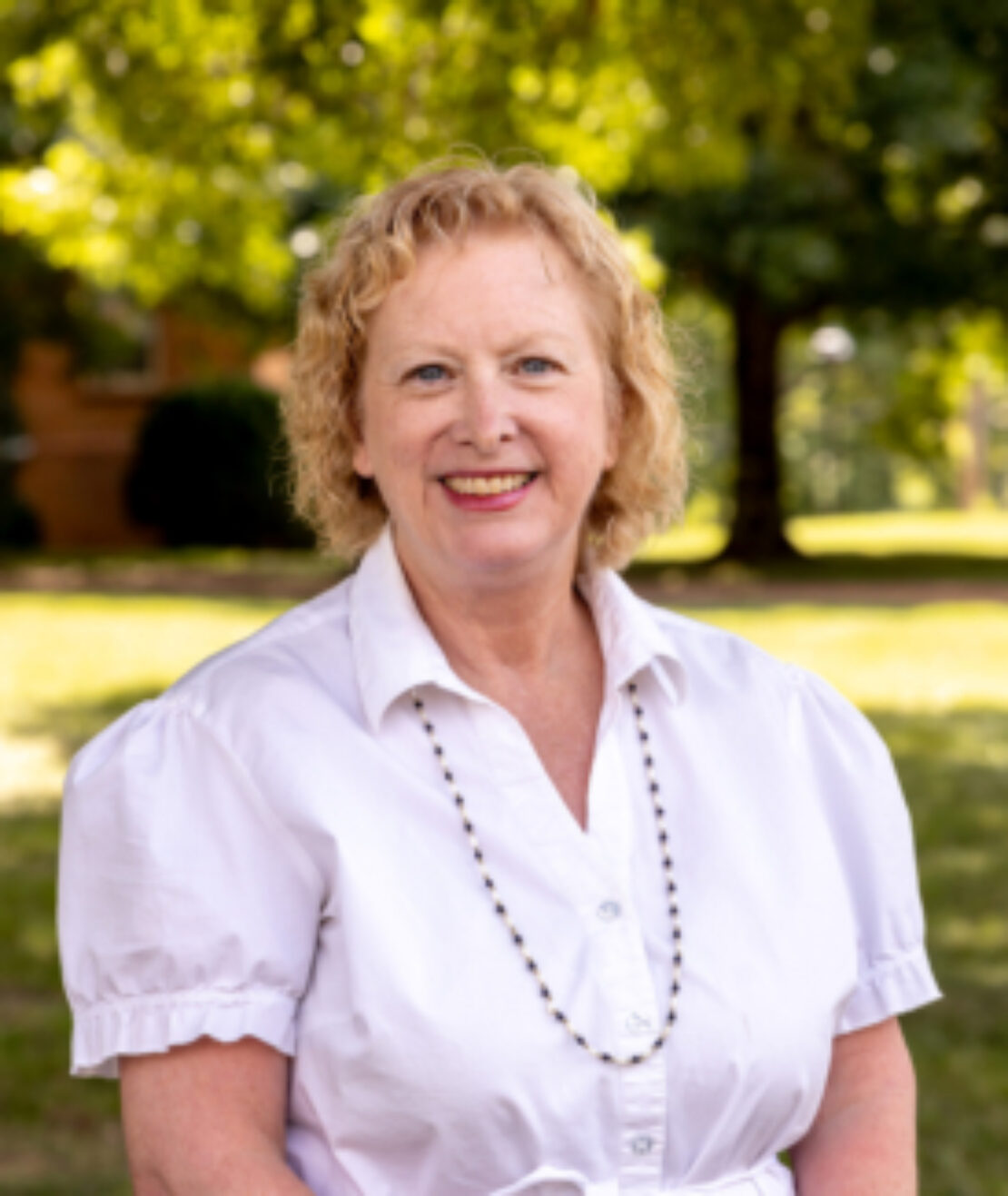 A smiling older woman with curly blonde hair stands outdoors, wearing a white short-sleeved blouse and a long beaded necklace. Green trees and grass are visible in the sunlit background.