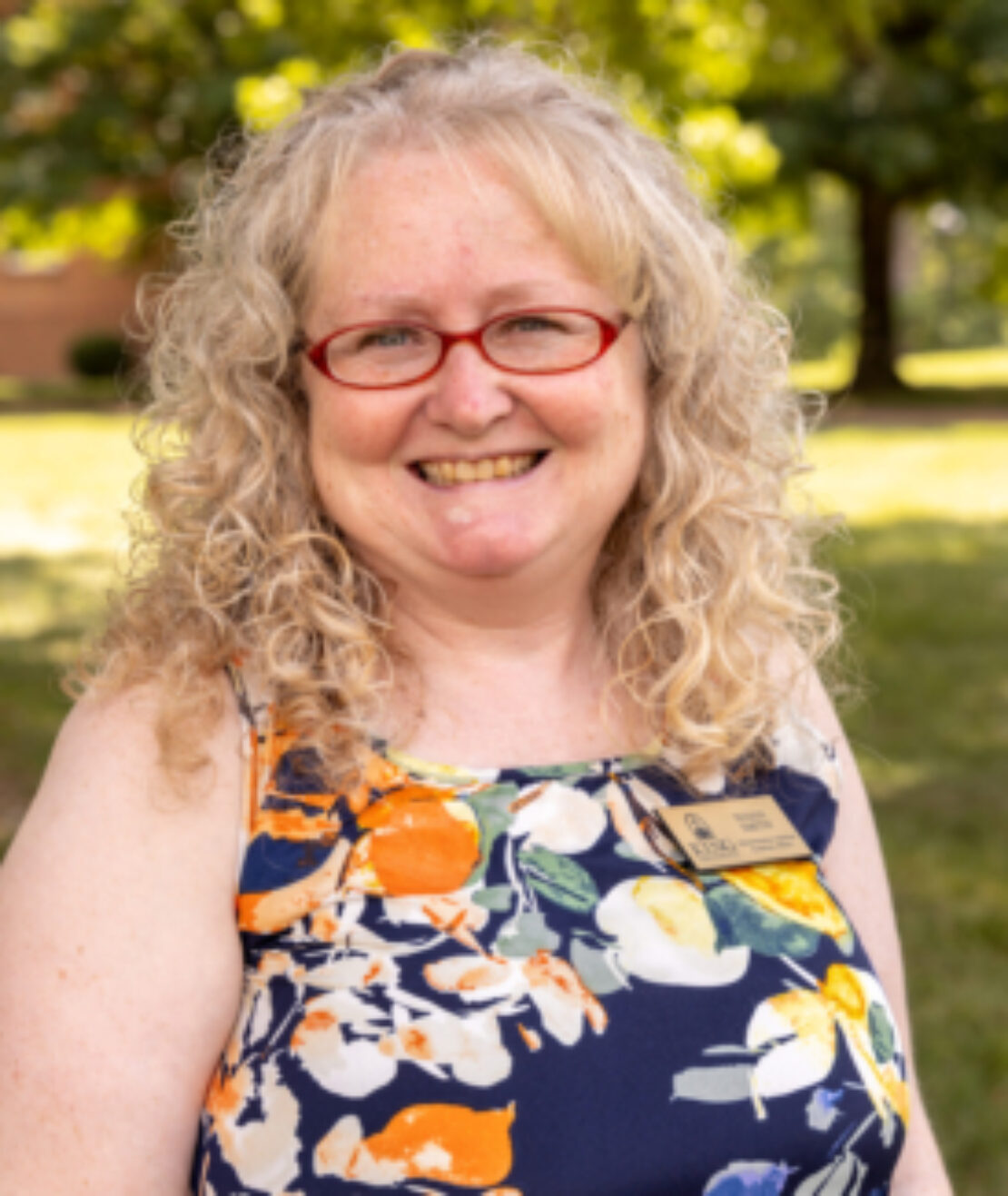 A woman with curly blonde hair and red glasses smiles outdoors. She is wearing a colorful floral dress and a name badge. The background is green and sunny with trees and grass.