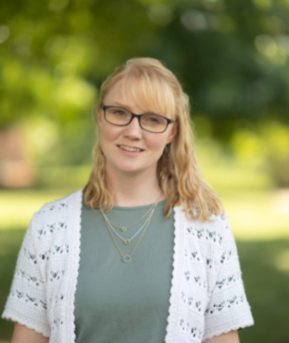 A woman with blonde hair and glasses stands outdoors, smiling. She wears a green top, a white knit cardigan, and layered necklaces. The background is blurred greenery, suggesting a park or garden setting.