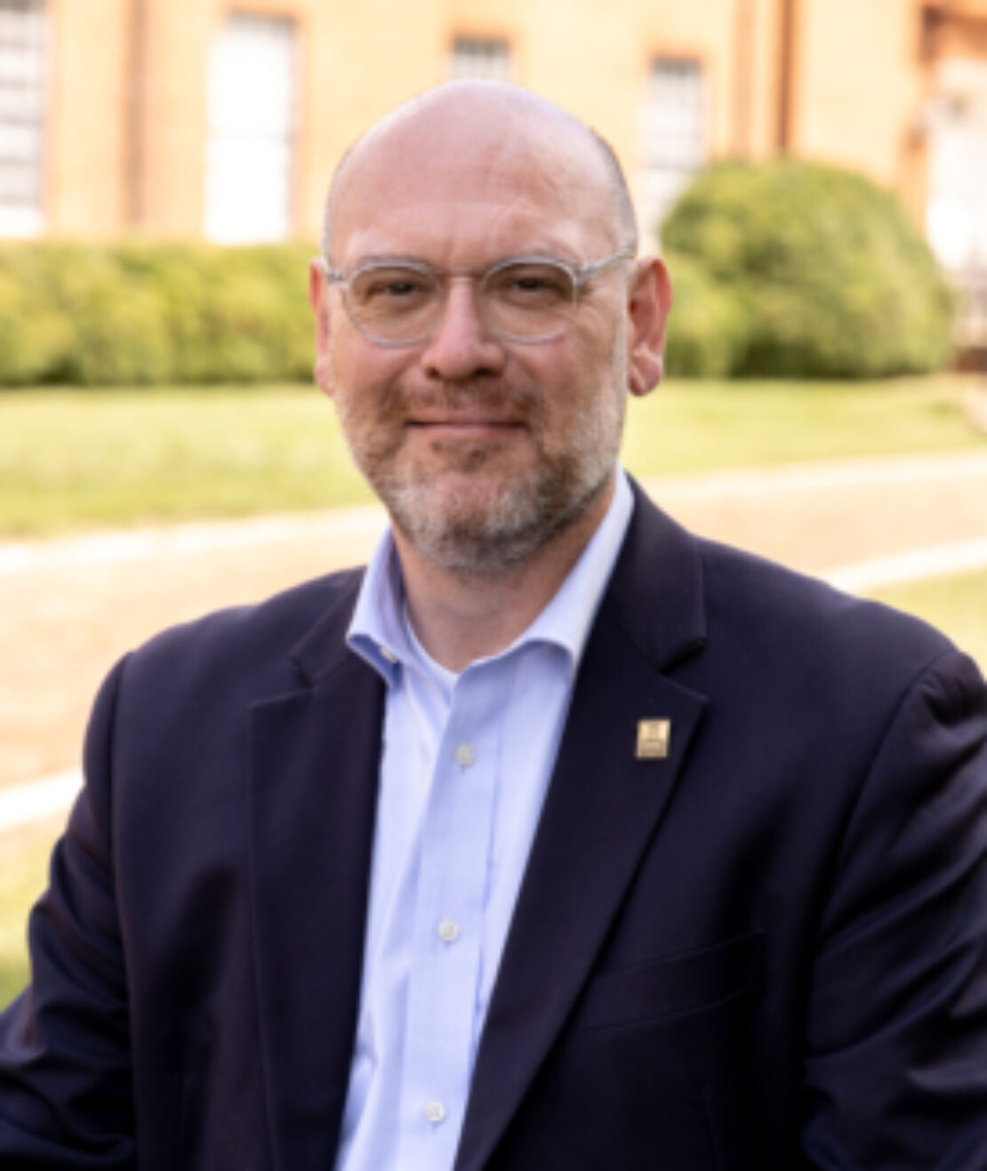 A man with glasses and a beard wearing a dark suit jacket and light blue shirt smiles outdoors in front of a brick building and green bushes.
