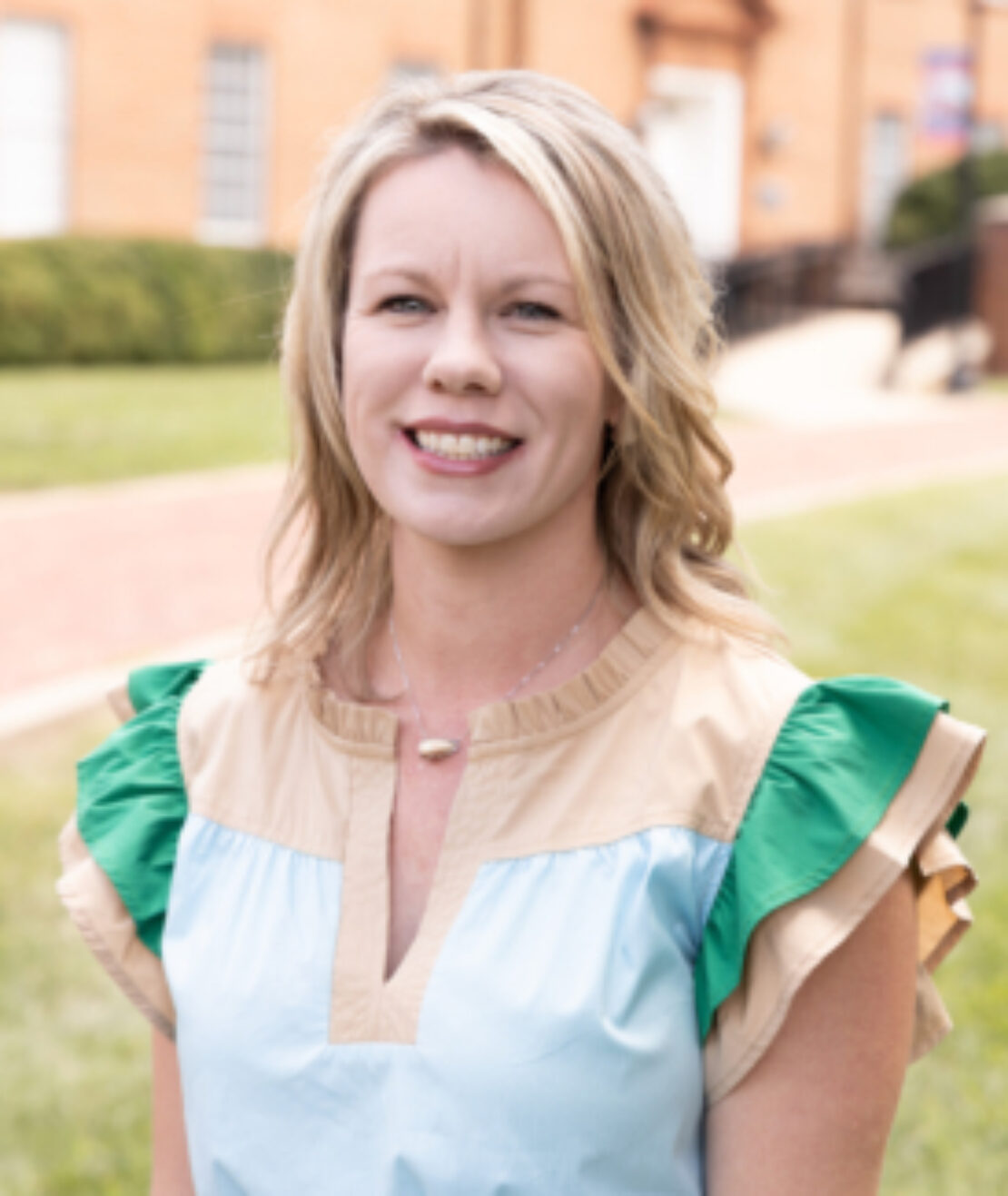 A woman with blonde hair wearing a light blue dress with tan and green ruffled sleeves smiles while standing outside in front of a brick building.