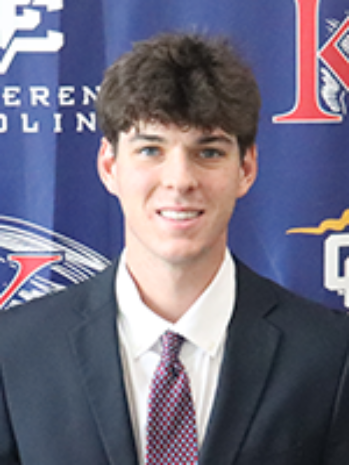 A young man with short brown hair wearing a dark suit, white shirt, and patterned tie stands in front of a blue backdrop with athletic logos and text.