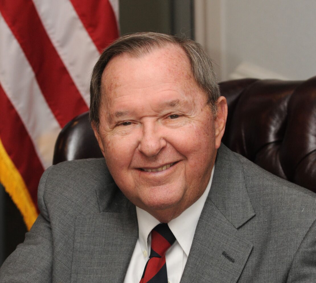 An older man in a gray suit and striped tie smiles while sitting in a leather chair. An American flag is partially visible in the background.