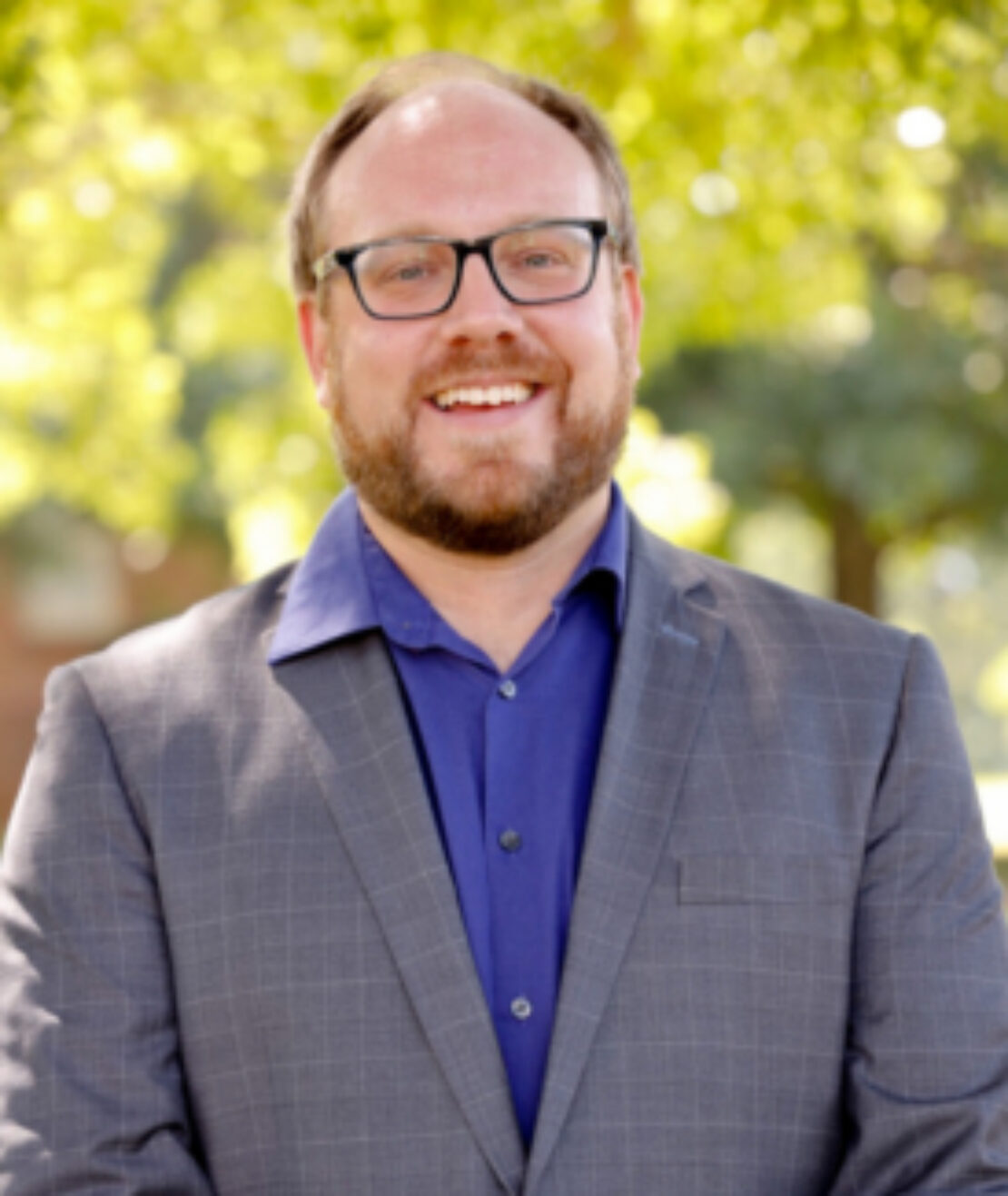 A smiling man with glasses and a beard wears a gray plaid suit jacket over a blue shirt. He stands outdoors in front of blurred green trees on a sunny day.