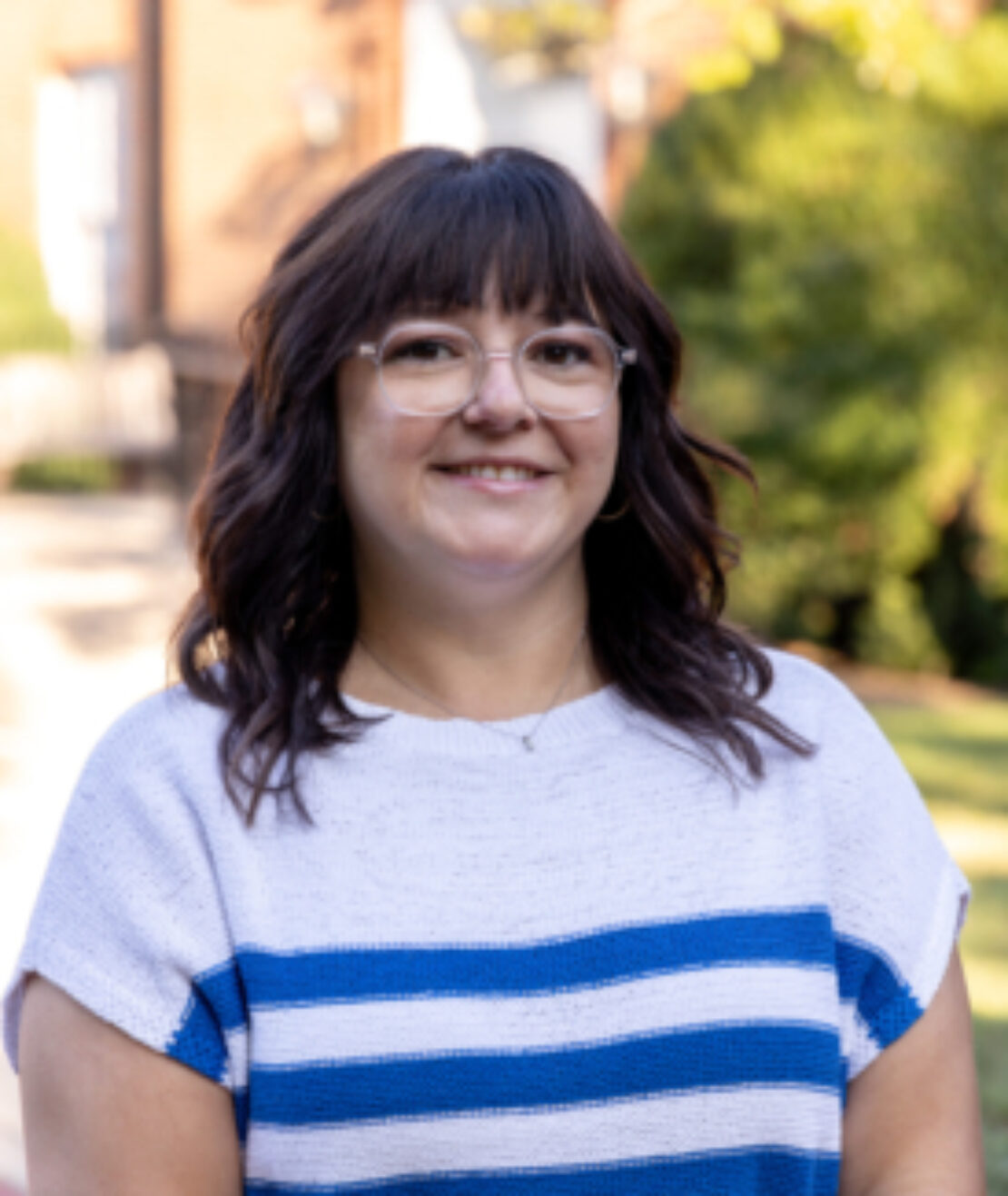 A woman with medium-length dark hair and glasses smiles outdoors. She is wearing a white sweater with blue stripes. Greenery and a blurred building are visible in the background.