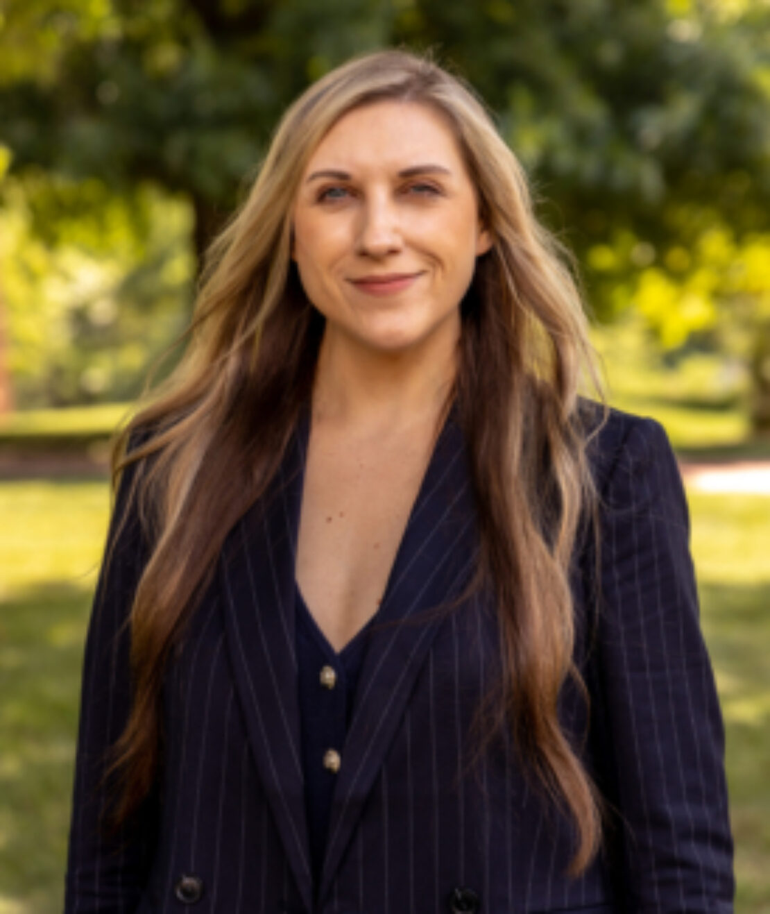 A woman with long, wavy blonde hair wearing a dark pinstripe blazer stands outside in a sunlit park with green trees and grass in the background, smiling at the camera.