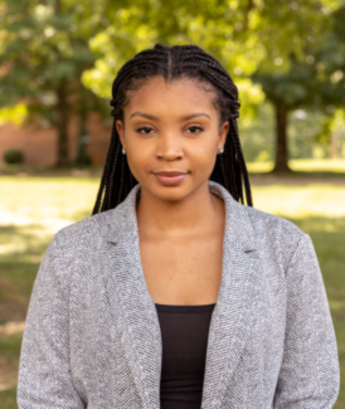 A young woman with braided hair, wearing a grey blazer and black top, stands outdoors on a sunny day with trees and greenery in the background.