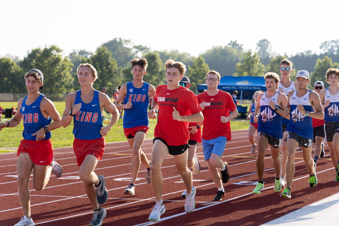 A group of teenage boys in athletic uniforms are running on a track outdoors during a race, with trees and a blue tent visible in the background. The sun is shining and the competitors look focused.