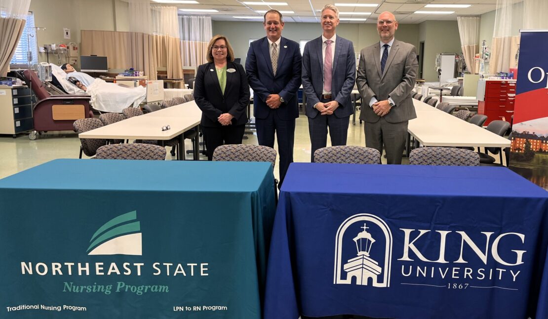 Four people in business attire stand behind tables with Northeast State Nursing Program and King University tablecloths in a classroom or medical training room, with medical equipment and beds visible in the background.