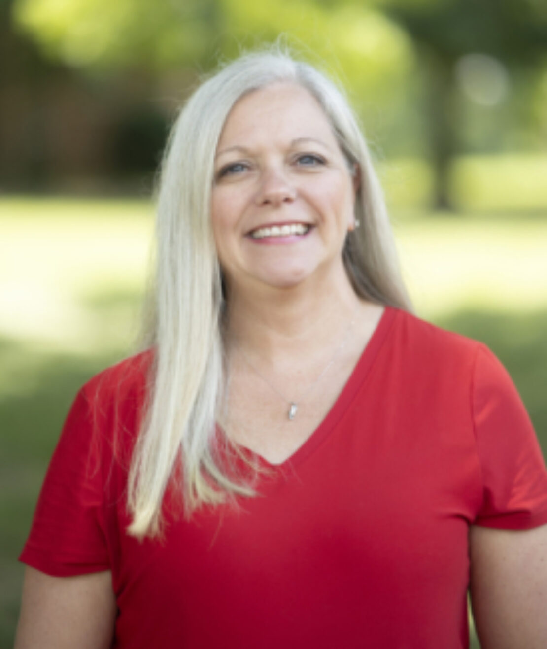 A smiling woman with long gray hair wearing a red shirt stands outdoors with soft green trees and sunlight in the background.