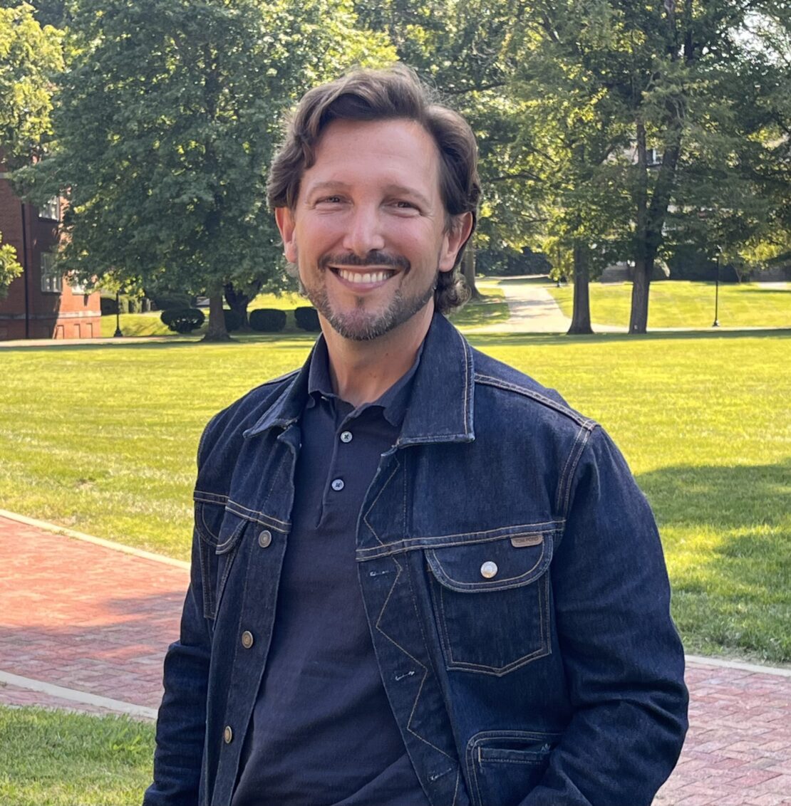 A smiling man with wavy brown hair and a trimmed beard stands on a brick path in a sunny, green park, wearing a dark denim jacket over a navy shirt. Trees and grassy areas are visible in the background.
