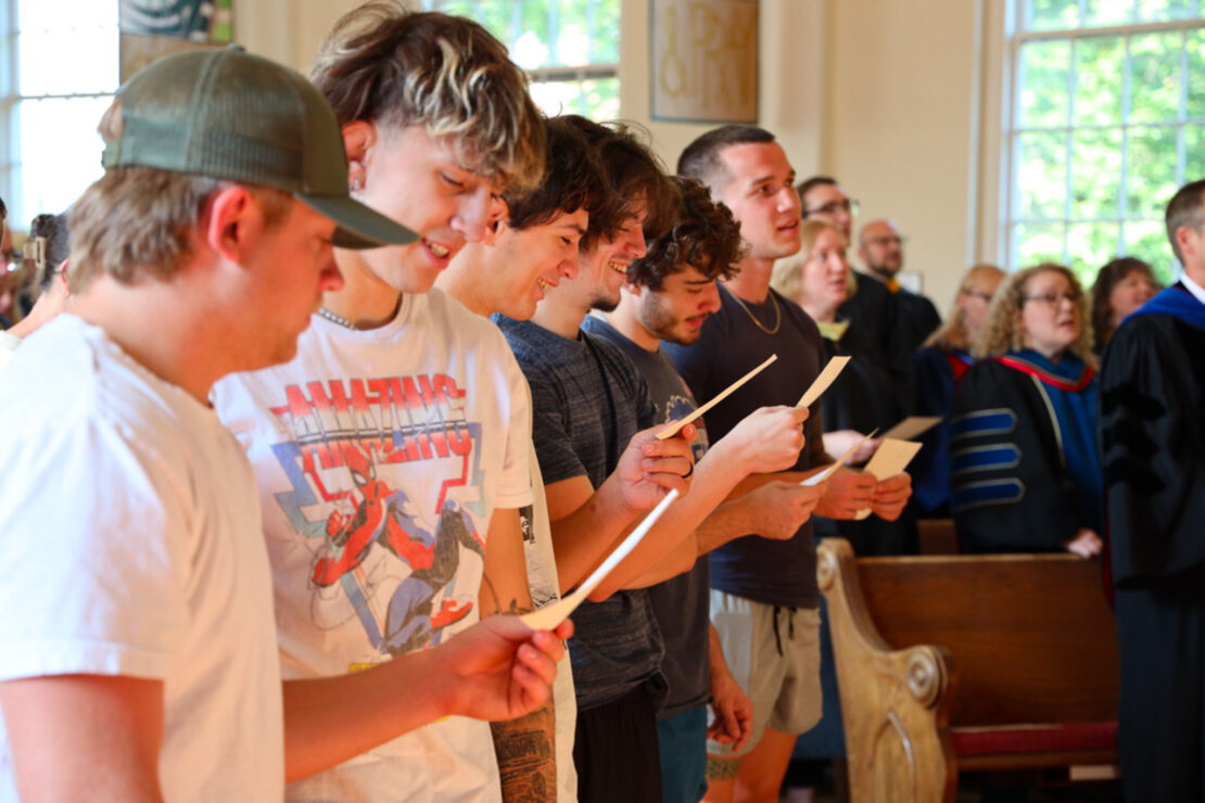 A group of young men standing in a row inside a building, holding papers and smiling, while people in academic robes and others sit in pews behind them. Sunlight streams through large windows in the background.