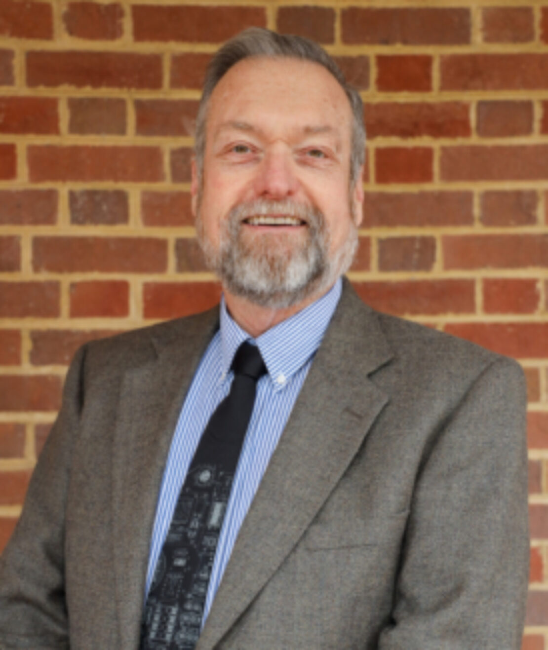 A middle-aged man with gray hair and a beard, wearing a brown blazer, striped shirt, and dark tie, stands smiling in front of a red brick wall.