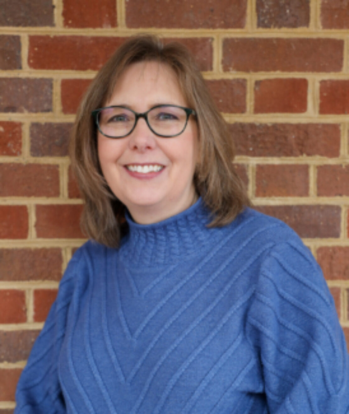 A woman with shoulder-length brown hair and glasses smiles while standing in front of a brick wall, wearing a textured blue sweater.