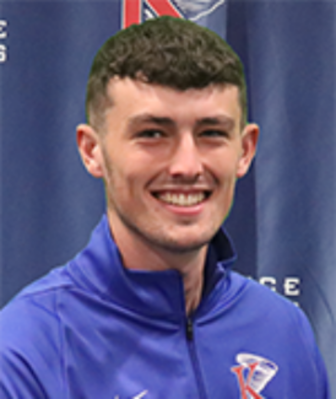 A young man with short brown hair smiles at the camera, wearing a royal blue athletic jacket with a K logo on the chest. The background features a blue banner with white text and similar K logos.