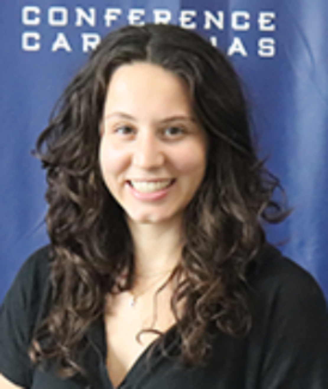 A woman with long, curly brown hair smiles at the camera. She is wearing a black top and is standing in front of a blue background with white text that is partially visible.