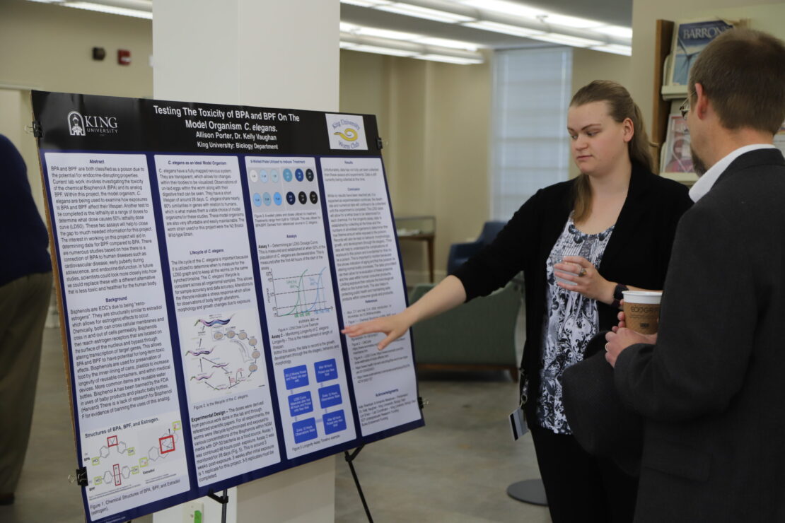 A woman explains a scientific research poster to a man holding a coffee cup in a well-lit indoor space. The poster contains text, charts, and diagrams about toxicity testing.