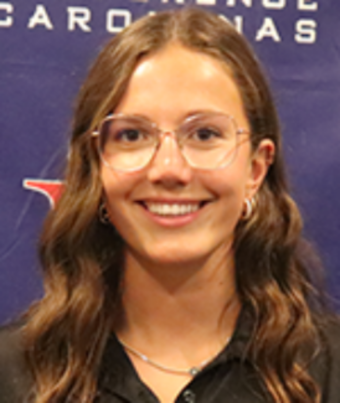 A young woman with long wavy brown hair and glasses smiles at the camera. She is wearing a black collared shirt and stands in front of a blue backdrop with white and red text and logos.
