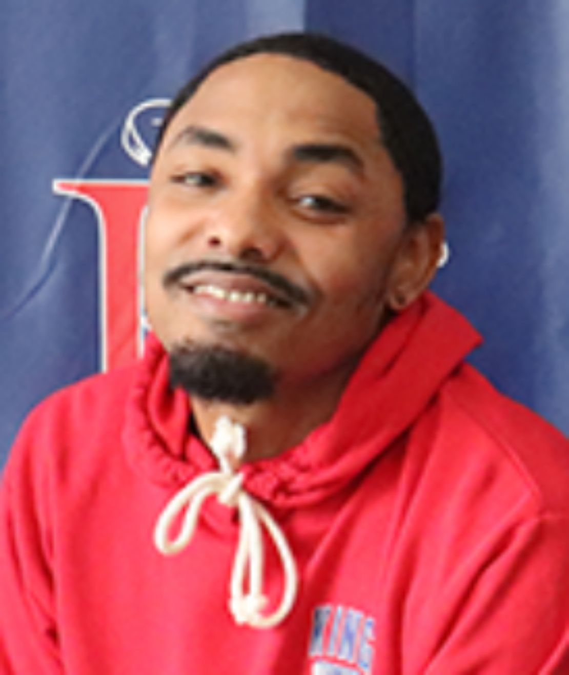 A man wearing a red hoodie with white drawstrings is smiling in front of a blue background with red and white lettering.