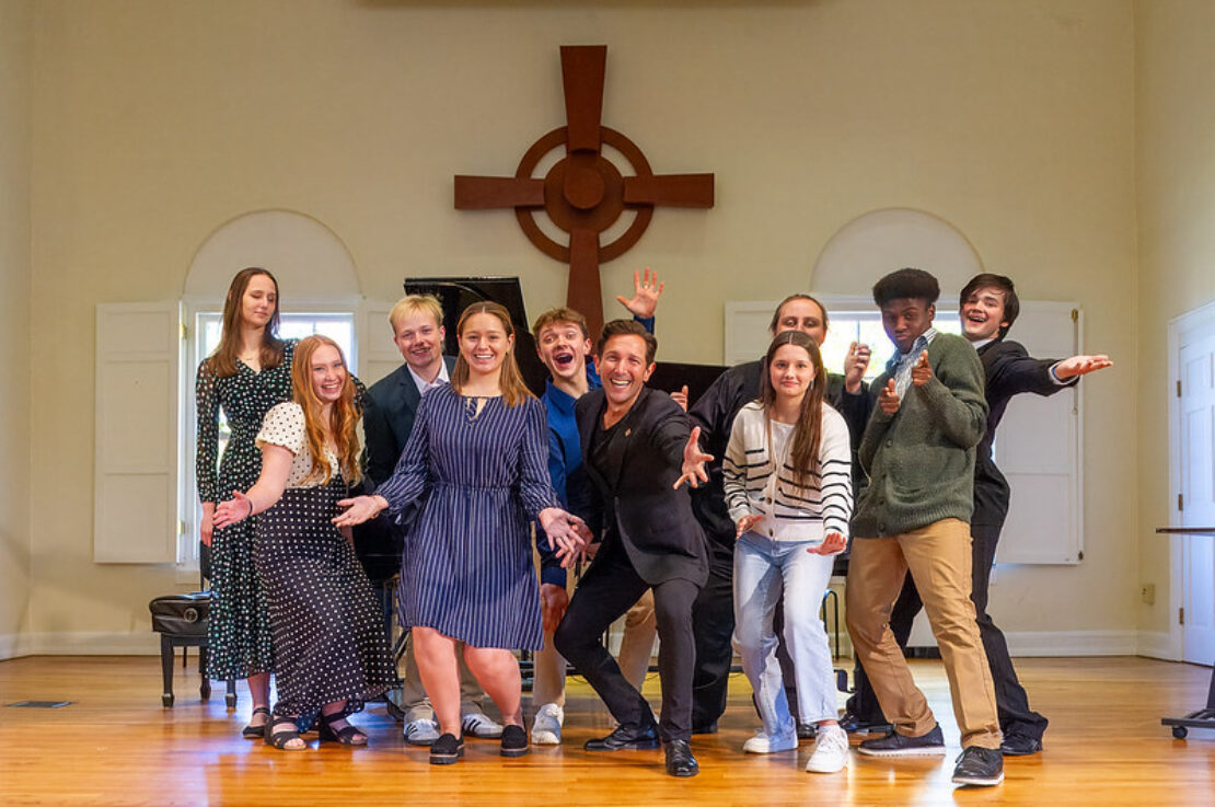 A group of ten young adults poses enthusiastically in a bright room with a wooden cross on the wall behind them. They are smiling, gesturing playfully, and appear to be celebrating or performing.
