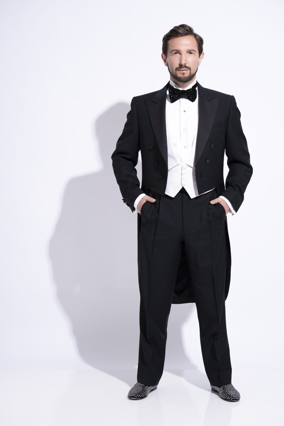 A man stands against a white background wearing a black tuxedo with tails, a white waistcoat, white shirt, black bow tie with white dots, and patterned shoes, with his hands in his pockets and looking confidently ahead.