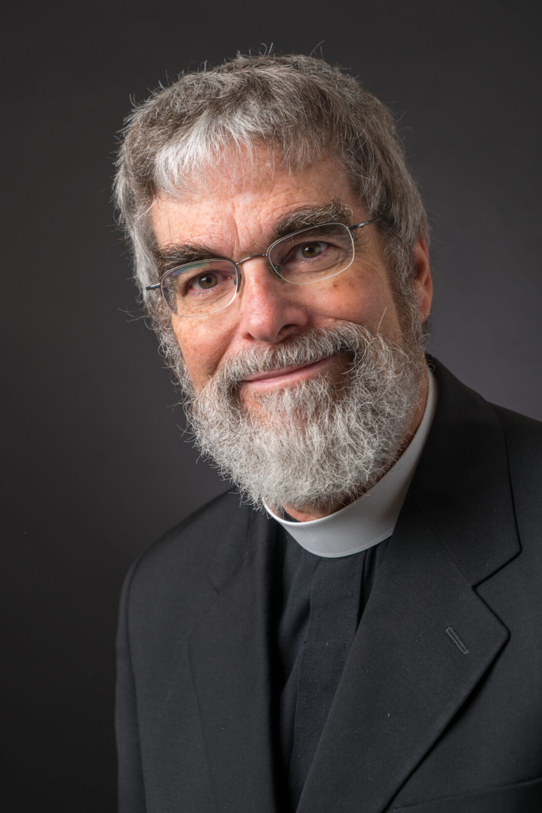 An older man with gray hair, glasses, and a full beard smiles while wearing a black suit and a clerical collar, posed against a dark background.