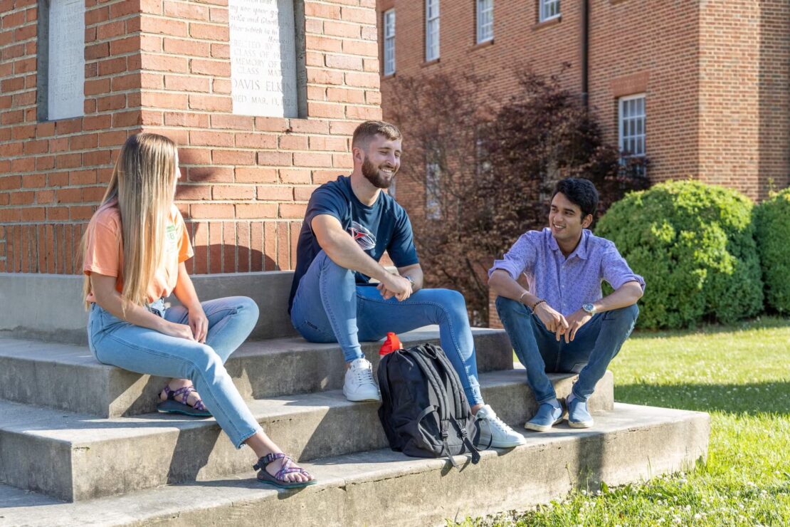 Three college students sit on brick steps outside, talking and smiling on a sunny day. Two men and one woman are casually dressed, with backpacks nearby and a grassy lawn and brick buildings in the background.