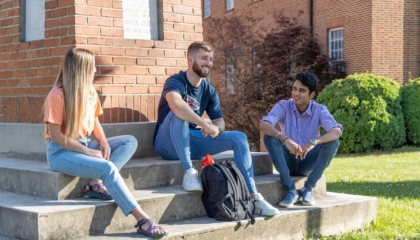 Three college students sit on brick steps outside, talking and smiling on a sunny day. Two men and one woman are casually dressed, with backpacks nearby and a grassy lawn and brick buildings in the background.