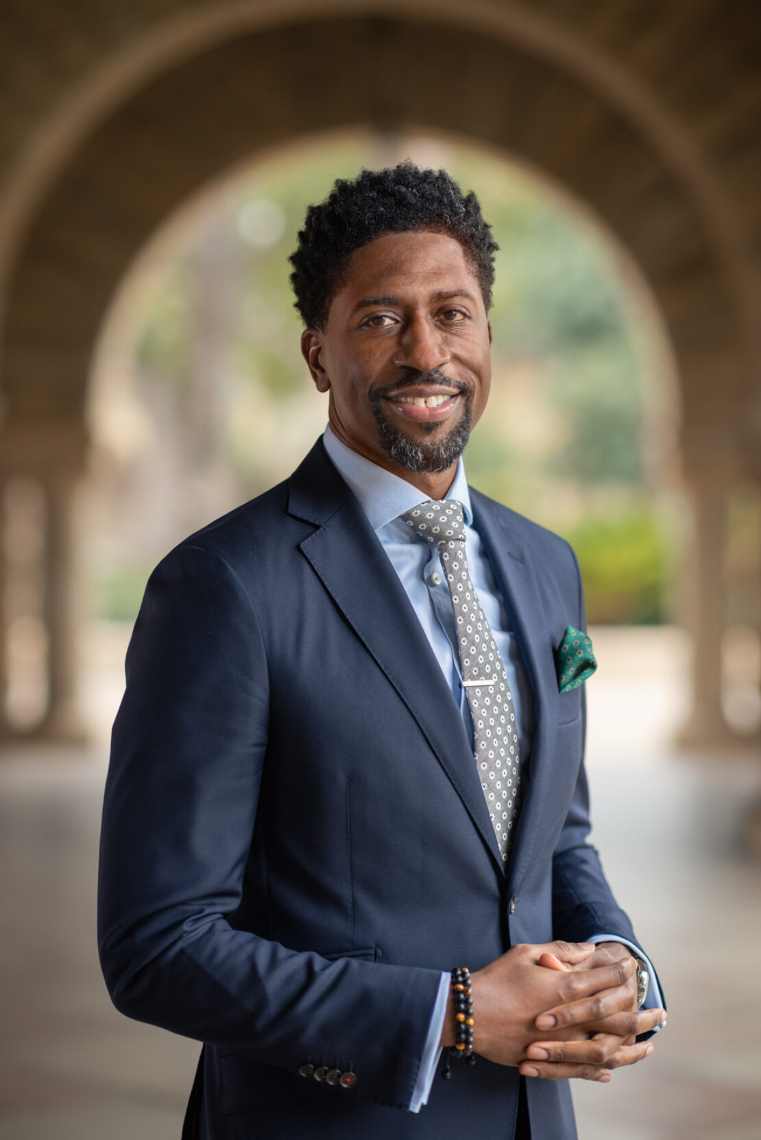A man in a navy suit with a patterned tie and pocket square stands smiling outdoors under an arched walkway, with his hands clasped in front of him.