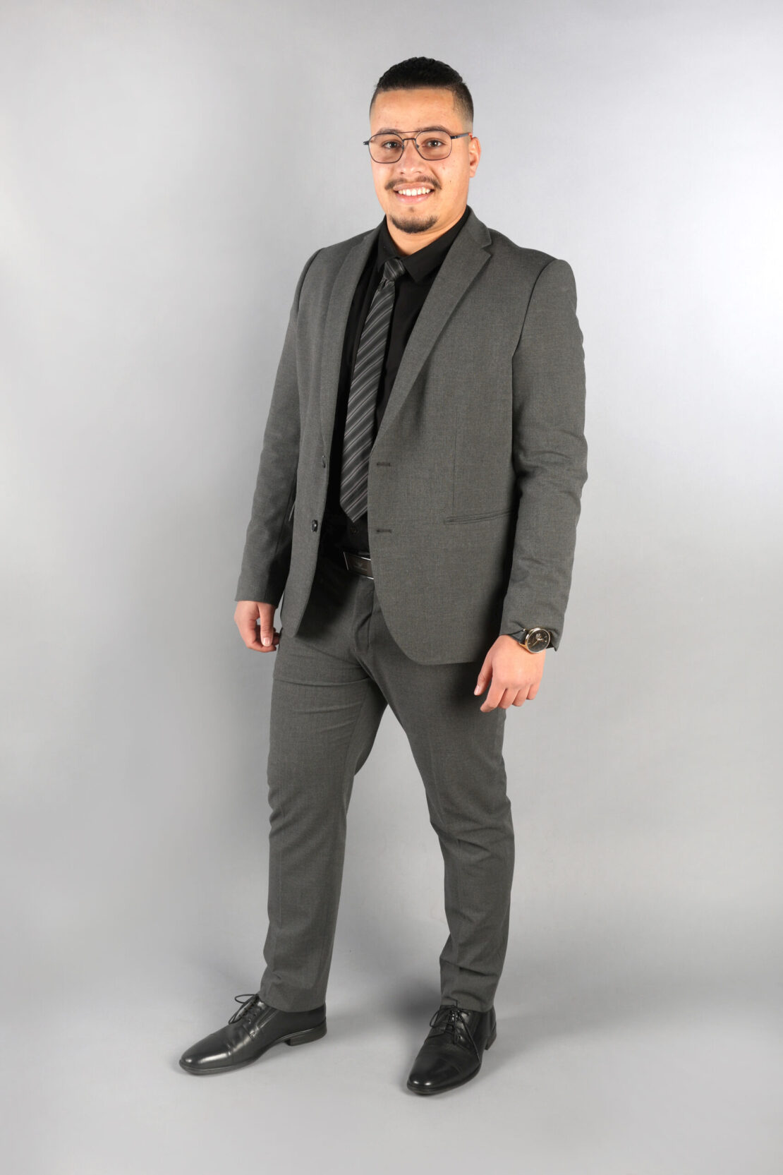 A man wearing glasses and a gray suit with a black shirt and striped tie stands smiling against a plain light gray background.