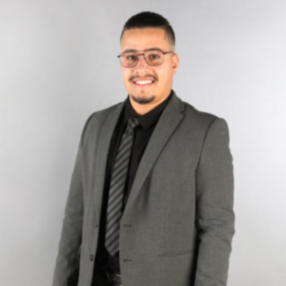 A man with short dark hair, glasses, and a mustache is wearing a gray suit, black shirt, and striped tie. He is smiling and standing against a plain light gray background.