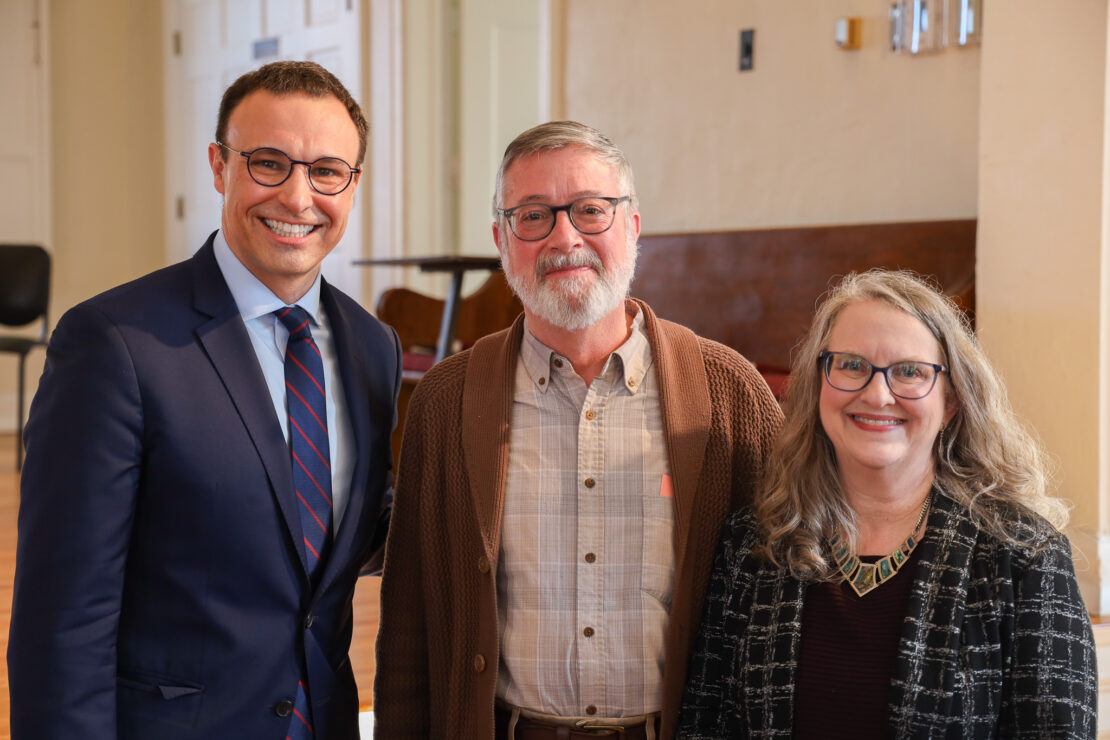 Three adults stand indoors, smiling at the camera. The man on the left wears a blue suit and glasses, the man in the middle wears a brown cardigan, and the woman on the right has long gray hair and glasses.
