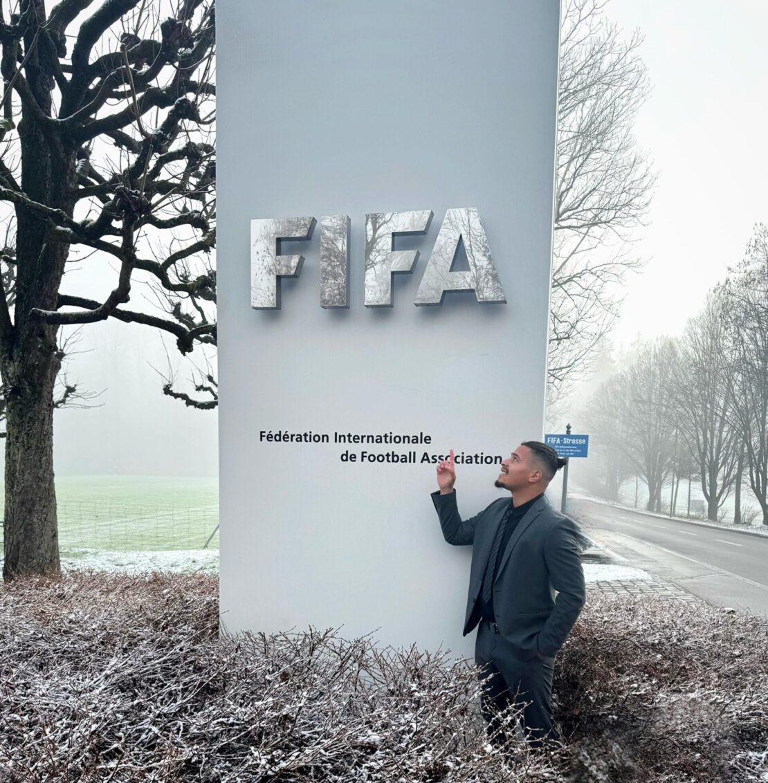A man in a suit stands beside a large vertical sign that reads FIFA Fédération Internationale de Football Association outdoors on a frosty day, with bare trees and a road in the background.