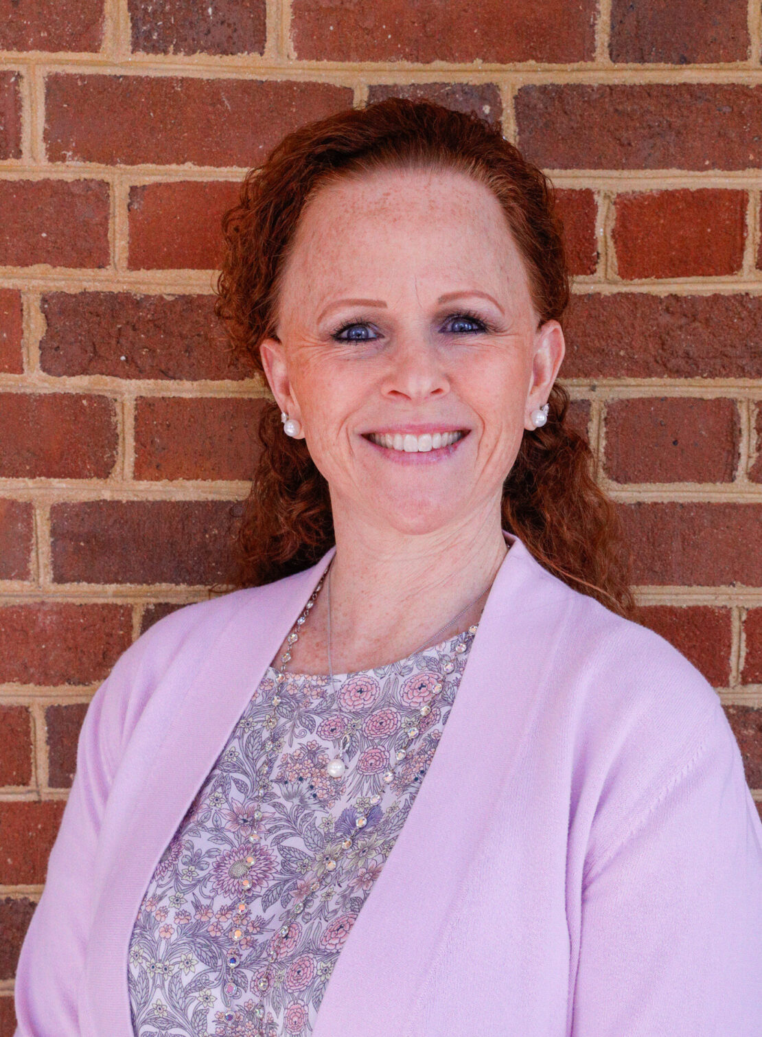 A woman with curly red hair, wearing a light pink cardigan and a floral top, smiles while standing in front of a brick wall.