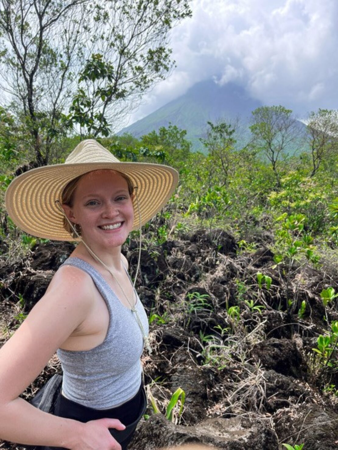A smiling woman in a straw hat and gray tank top stands outdoors on rocky terrain, surrounded by lush greenery, with a cloud-covered volcano in the background.