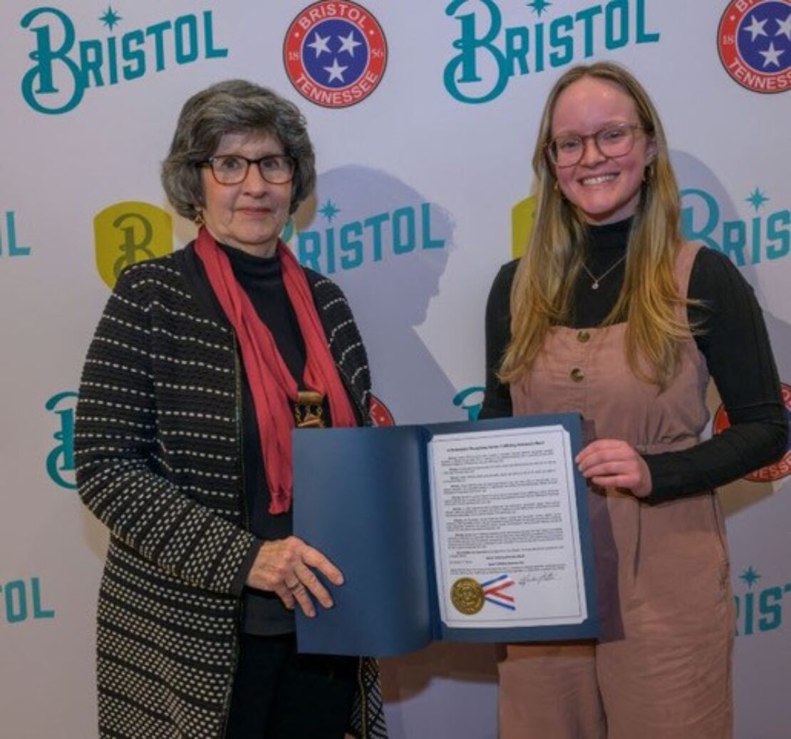 Two women stand together as one holds an open blue folder displaying an official document with a gold seal in front of a backdrop featuring “Bristol” and the Bristol, Tennessee logo.