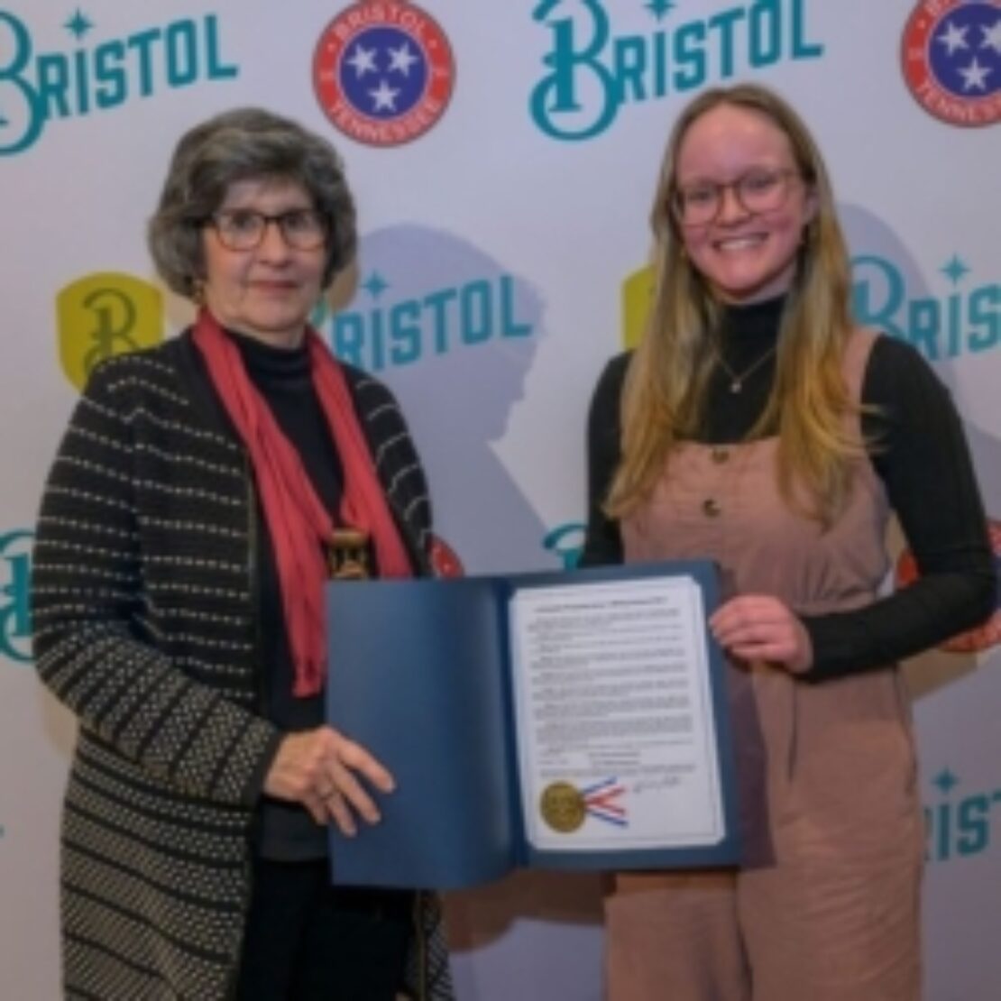 Two women stand together smiling; one holds an open folder displaying a document. They are in front of a backdrop with Bristol, Tennessee, and related logos.